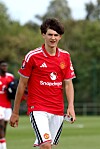Fotballspiller i rød Manchester United-drakt går på gresset, med lagkamerater og trær i bakgrunnen.